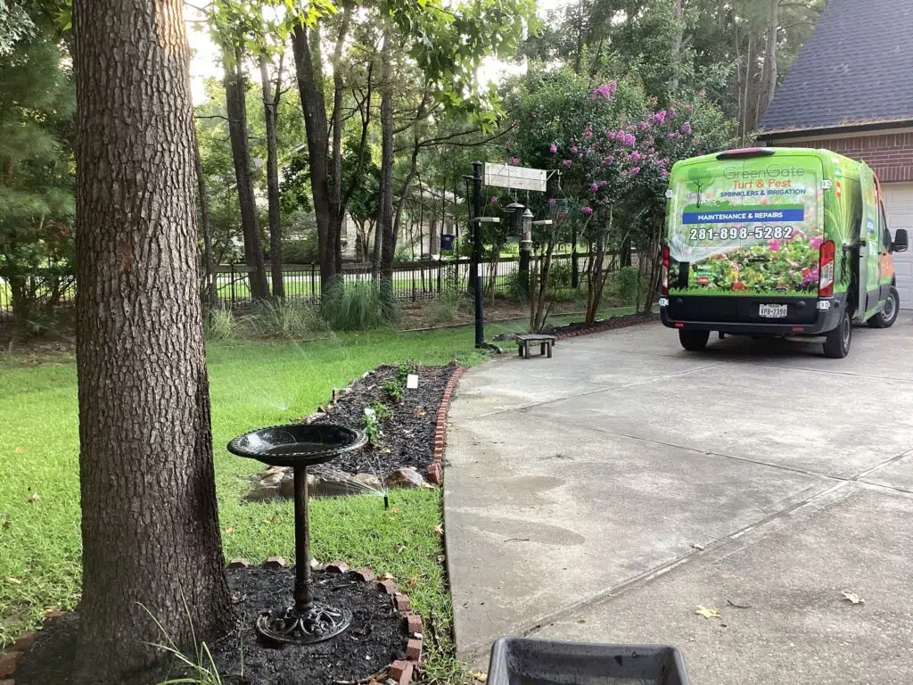 greengate van parked in a driveway with green grass and a flower bed to the left. Sprinklers are running during a sprinkler system maintenance service